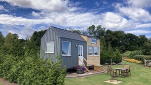 A small, two-story cabin with a grey exterior and a wooden upper floor, surrounded by lush greenery and a well-maintained lawn. - The Cedar Tiny House (Coldingham)