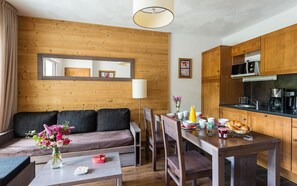 Un salon chaleureux avec un mur en bois, un canapé, une table à manger avec des chaises et un coin cuisine doté de meubles et d’appareils électroménagers en bois.