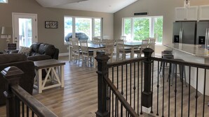 A spacious living room with a high ceiling and a dining area with white chairs and a table. There is a kitchen area with stainless steel appliances and a large window.