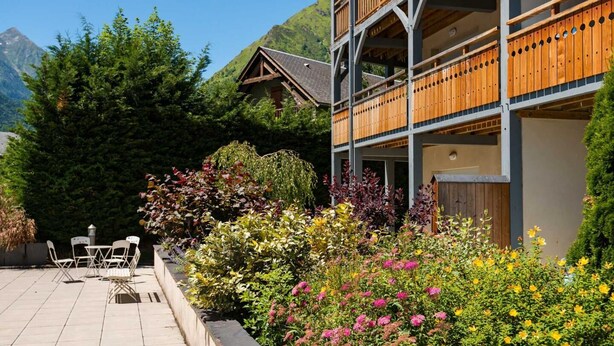 Kaksikerroksinen rakennus, jossa on puinen parveke ja puutarha, jossa kasvaa monenlaisia kasveja ja kukkia.