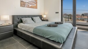 Een moderne slaapkamer met een groot bed, twee nachtkastjes, een schilderij aan de muur en uitzicht door een groot raam.