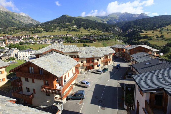 Vila montanhosa com edifícios de madeira tradicionais, uma área de estacionamento central e vista para as colinas ao redor.