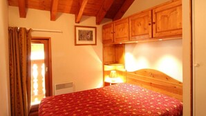Une chambre avec un plafond en bois, un lit recouvert d’une courtepointe à motifs floraux rouges, une table de chevet avec une lampe et des armoires en bois au-dessus du lit.