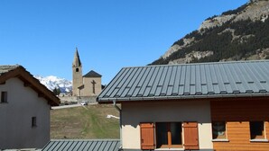 Un bâtiment avec un toit gris et des volets en bois, une église dotée d’un clocher, et un paysage montagneux en arrière-plan.