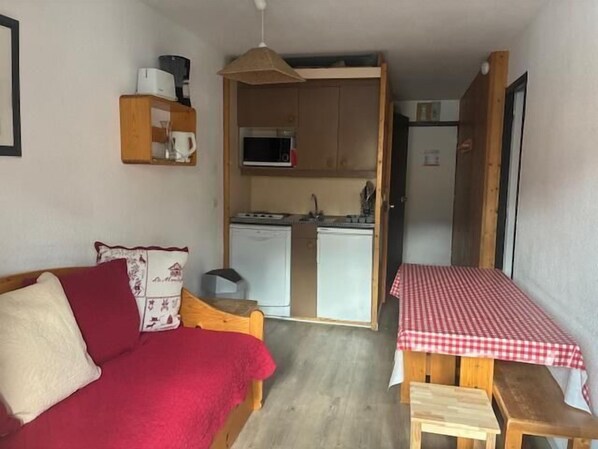 Una habitación compacta con un sofá rojo, una mesita de madera con mantel a cuadros y una kitchenette equipada con microondas y nevera.