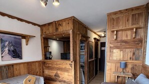 Een knus vertrek met een houten wand, een eettafel met stoelen, een bank en een klein keukentje.