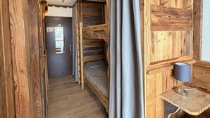 Een rustieke houten kamer met stapelbedden, een klein bureau met een lamp en een deur naar een andere kamer.