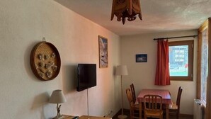 A room with a wooden desk, a television mounted on the wall, a dining table with chairs, a window with curtains, and a lamp.