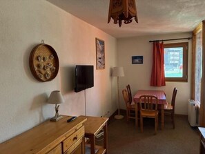 Quarto com escrivaninha de madeira, televisão na parede, mesa de jantar com cadeiras, janela com cortinas e luminária.