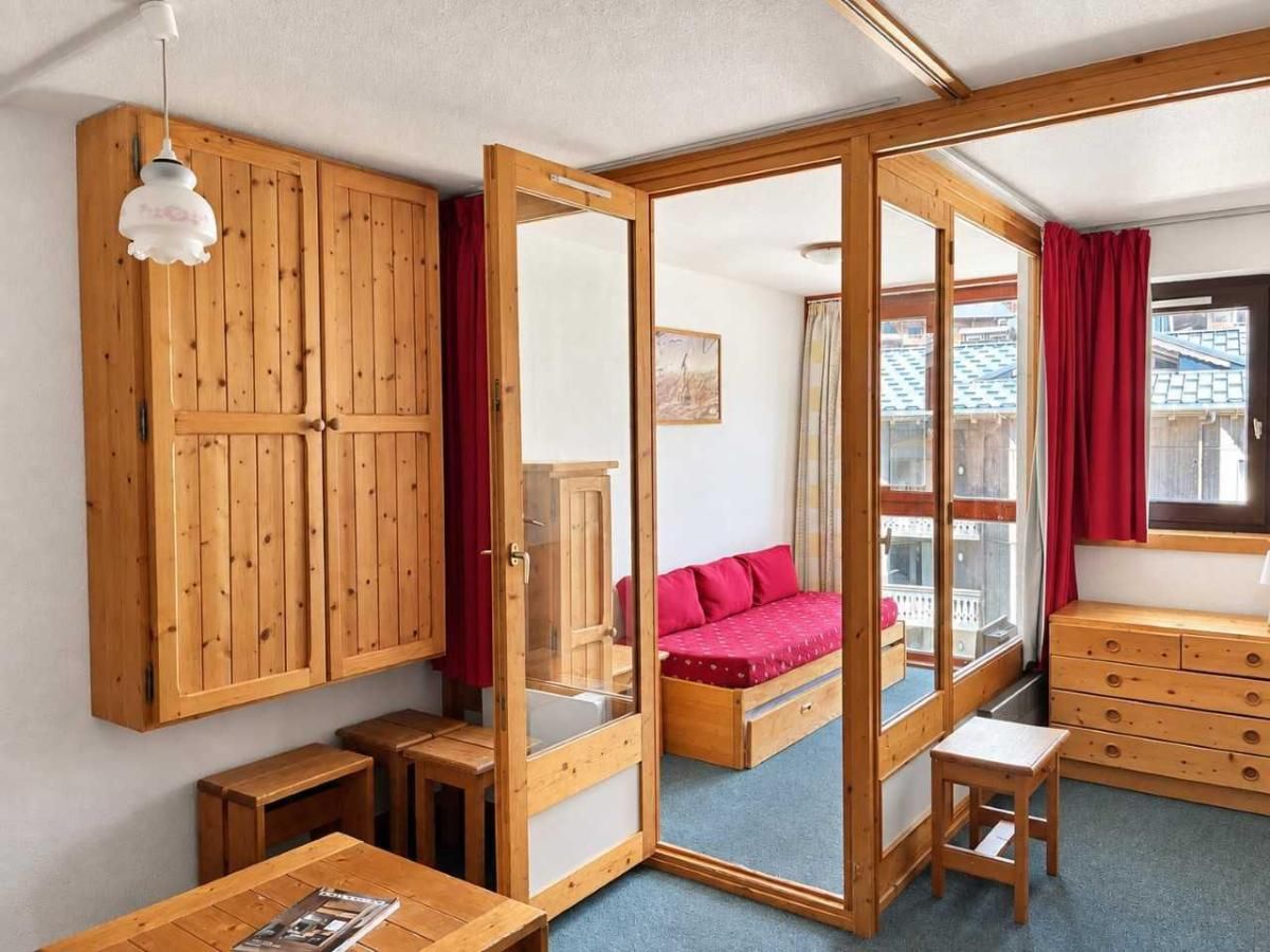 A room with a wooden wardrobe, a bed with red bedding, a wooden dresser, and a window with a view of buildings.