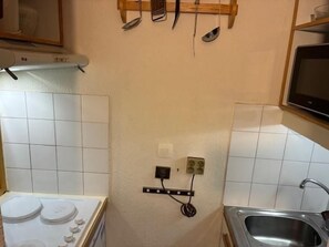 A small kitchen with white tiles, a stove, a sink, and a microwave. There are utensils hanging on the wall and a power outlet with a cord plugged in.