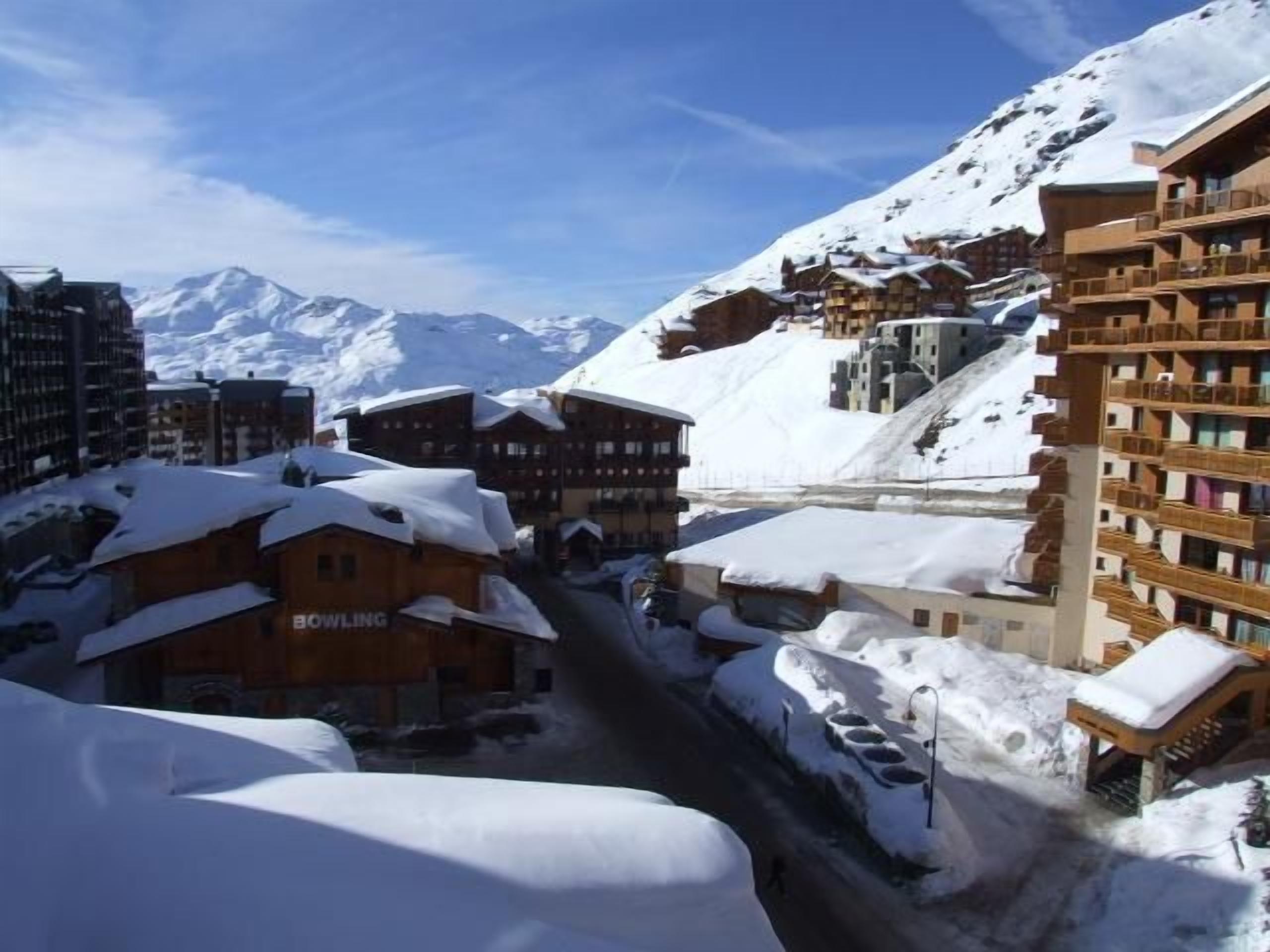 Een besneeuwde plaats in de bergen met gebouwen, waaronder een hotel genaamd 'BOWLING', en een skipiste op de achtergrond.