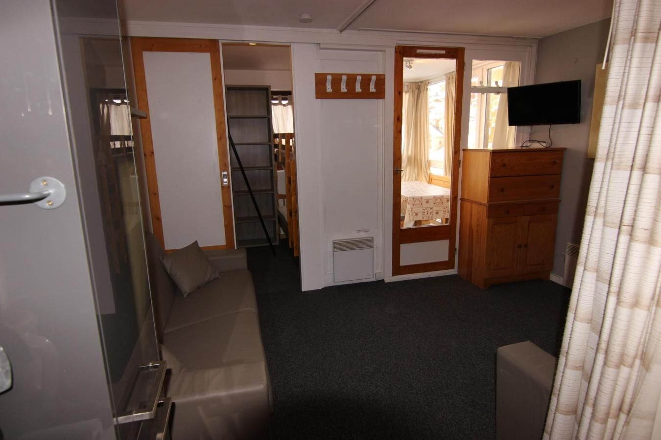 A compact room with a sofa, a wooden dresser, a television, and a door leading to another room.
