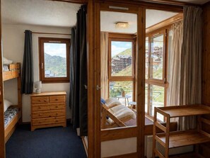 Ein kleines Zimmer mit einem Etagenbett, einem hölzernen Schminktisch und einem Fenster mit Blick auf Gebäude und Hügel.