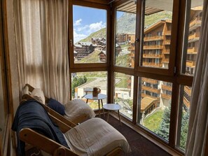 A room with a large window, two wooden beds, a small table, and a view of a mountainous area with buildings.