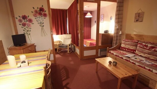 Ein gemütliches Zimmer mit Holztisch, einer Couch und einem Bett mit roten Vorhängen.