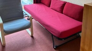 Ein Zimmer mit einem roten Sofa, einem grauen Sessel, einem Fenster mit Blick auf Gebäude und einem roten Vorhang.