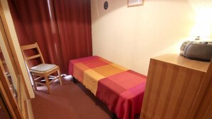 Ein kleines Zimmer mit einem Einzelbett, einem Stuhl, einem Nachttisch aus Holz und einer Lampe.