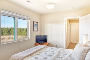 A bedroom with a bed, a TV on a stand, a window with a view of greenery, and a door leading to another room.