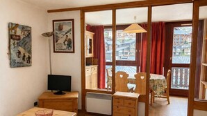 A room with a wooden desk, a television, a radiator, a window with red curtains, and a view of buildings outside. - 50m from ski slopes, 6th floor, balcony, tv, 35m², Val Thorens (Val Thorens)