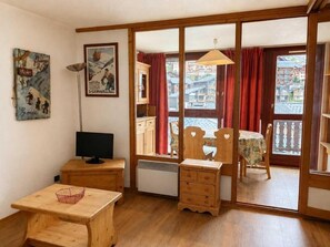 Ein Zimmer mit einem Holztisch, einem Fernseher, einer Heizung, einem Fenster mit roten Vorhängen und Blick auf Gebäude.