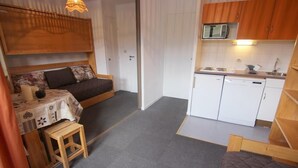 Une petite pièce comprenant un lit, une petite table, un tabouret, un coin cuisine et une porte donnant sur une autre pièce.