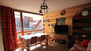 Een gezellige woonkamer met een eethoek, houten meubels, een televisie en uitzicht op gebouwen.