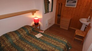 Een slaapkamer met een bed, nachtkastje met lamp, houten stoel, en een badkamer met wastafel en spiegel.