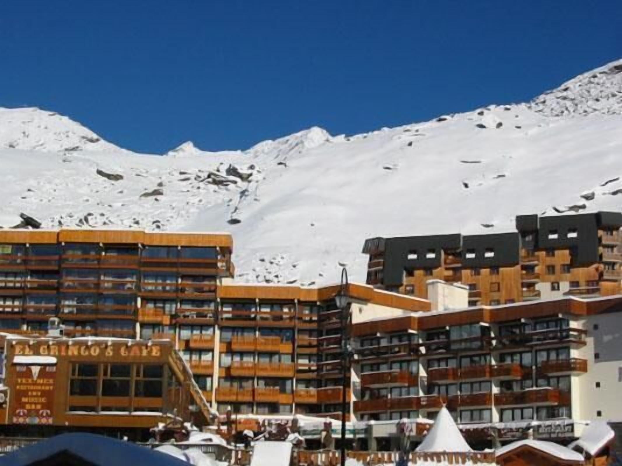 Un hotel de varios pisos, con una estación de esquí al fondo y un cielo azul despejado.