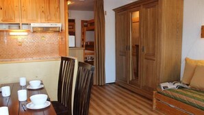 Een compacte kamer met een houten eettafel, stoelen, een bed met een groen en beige dekbedovertrek en een houten garderobe.