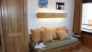 Een houten bed met een gewatteerde groene spreideken en meerdere kussens, een houten garderobe en een wandlamp.