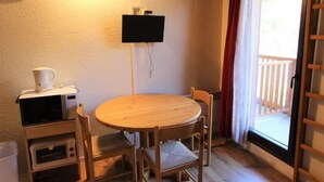 Una habitación pequeña con una mesa redonda de madera y cuatro sillas, un televisor montado en la pared, y una ventana con vista a una terraza de madera.