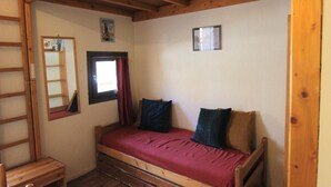 Una habitación pequeña con cama individual, techo de madera, ventana con cortinas rojas, espejo y una pequeña mesa de madera.