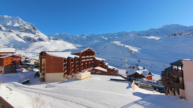 Un village de montagne enneigé, avec ses bâtiments traditionnels en bois, un télésiège et un ciel d’un bleu éclatant.