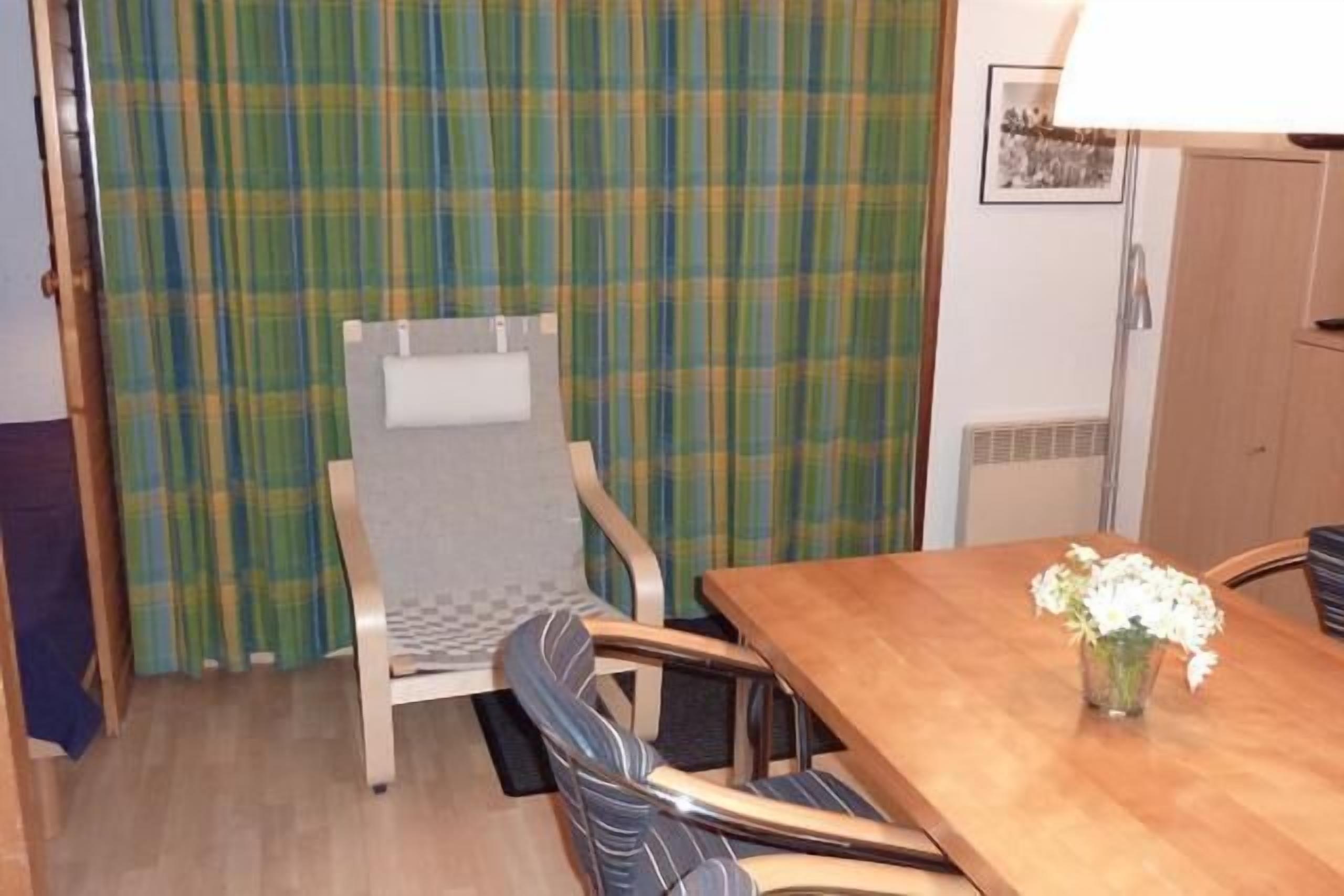 A dining area with a wooden table, two chairs, a plaid curtain, and a vase with flowers.