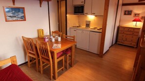 A cozy room with a wooden dining table, chairs, a red sofa, a kitchen with white cabinets, a microwave, and a red lamp. - 150m from ski slopes, floor -1, balcony, 29m², Val Thorens (Val Thorens)