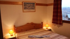 En pænt redt seng med et stribet tæppe, to sengeborde med lamper, et billede i ramme på væggen og et vindue med udsigt til en bygning.