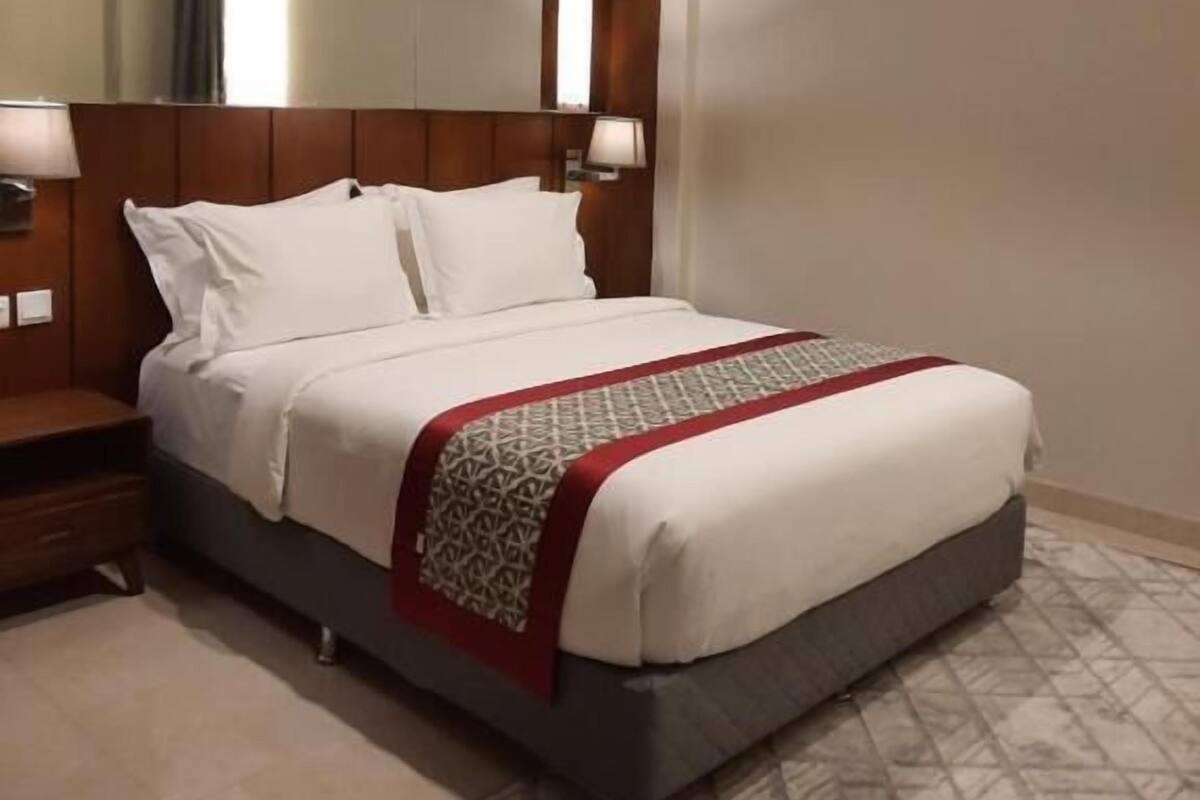 A hotel room with a large bed, a wooden headboard, two bedside lamps, a patterned throw blanket, and a tiled floor.