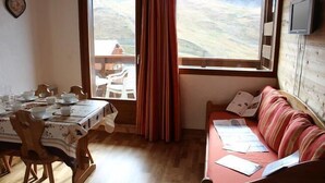 Una habitación acogedora con una mesa de comedor lista para cuatro personas, un sofá con papeles y una ventana con cortinas.