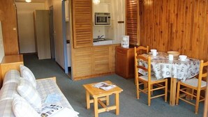 A compact room with a wooden wall, a kitchenette, a dining table with four chairs, a sofa, and a small wooden stool.