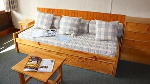A wooden bed with a checkered blanket and pillows, a wooden nightstand with a magazine, and a small wooden table with a magazine.