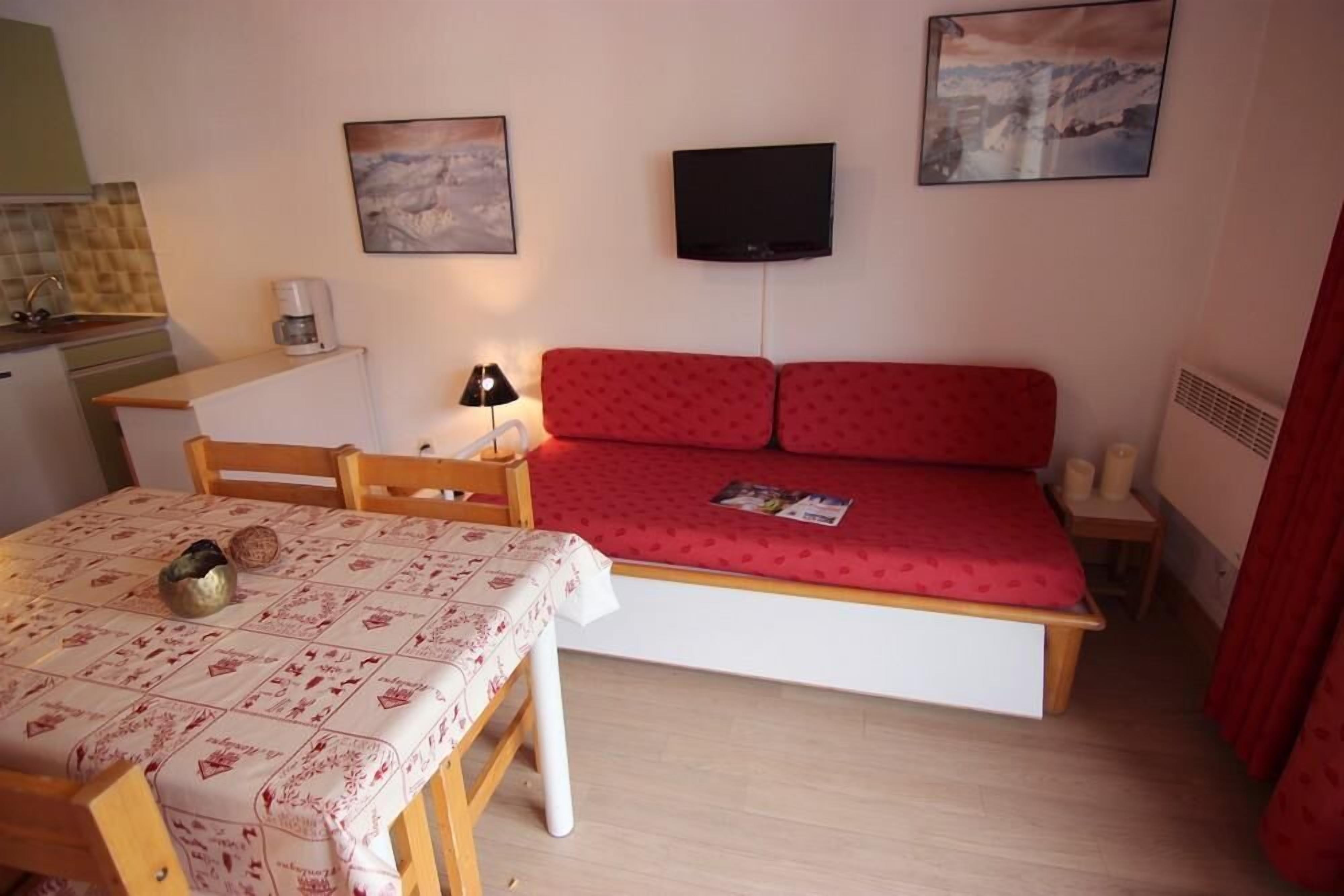 A compact room with a bed, a table with a tablecloth, a chair, a small table with a lamp, and a television on the wall.