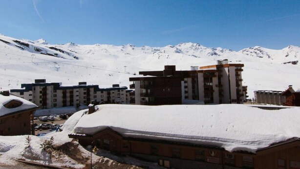 Complejo de edificios cubierto de nieve con varios pisos, probablemente un hotel, con montañas nevadas al fondo bajo un cielo despejado.