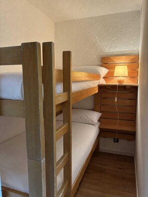 A bunk bed with a wooden ladder, a bedside table with a lamp, and a wall-mounted shelf.