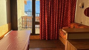 A room with a bed, a small table, and a view of buildings and mountains through a window. - 200m from resort center, 2nd floor, balcony, tv, 21m², Val Thorens (Val Thorens)