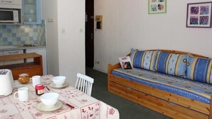 Een kleine, gezellige kamer met een kitchenette, een houten bank, een eettafel met schalen en een potje olijven, en een muur met ingelijste foto’s.