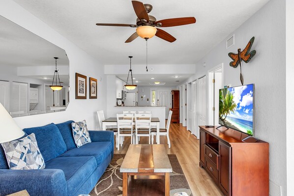 A modern living room with a blue sofa, wooden coffee table, ceiling fan, TV, and kitchen in the background.