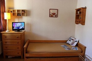 Una habitación pequeña y sencilla con una cama de madera, una mesita de noche también de madera con una lámpara, un televisor y una estantería con objetos decorativos.