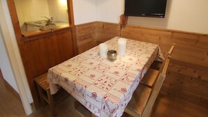 Una habitación pequeña con un banco y una mesa de madera, un televisor montado en la pared y una cocina visible a través de un separador de madera.