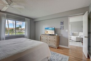 A bedroom with a bed, a television on a wicker console, a view of the outdoors through a window, and a door leading to another room. - Naples Getaway: Beach, Pool & Family-Friendly (Naples)